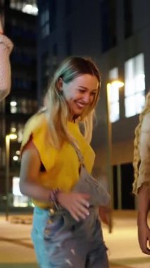 Dikey video. Mutlu genç kız arkadaşlar şehirde gece başarısını kutlamak için dans ediyorlar. Sadece kadınların birlikte dışarıda eğlendiği gülümseyen kadın grubu. Çok kültürlü insanların neşeli topluluğu.