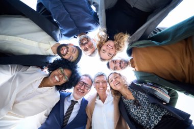 Çeşitli iş adamlarından oluşan neşeli bir grubun düşük açılı görüntüsü resmi bir toplulukta toplanıyor, gülümsemeleri paylaşıyor ve resmi bir etkinlikte birlik ve kutlama başarısı göstergesi olarak birbirlerine sarılıyorlar.