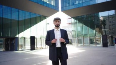 Beyaz tenli, şık mavi takım elbiseli, şapkalı ve gözlüklü genç bir hippi bir ofis binasının önünde ciddi ve kendinden emin bir şekilde duruyor. Ceketini resmi bir iş etkinliğine hazırlıyor.