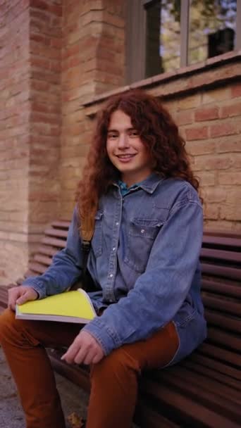 カメラを見ている大学ビル外の若い幸せな白人赤い頭の学生の垂直の肖像画 ベンチの屋外に座っている彼女の手のフォルダを保持しているGen Z女性 — ストック動画