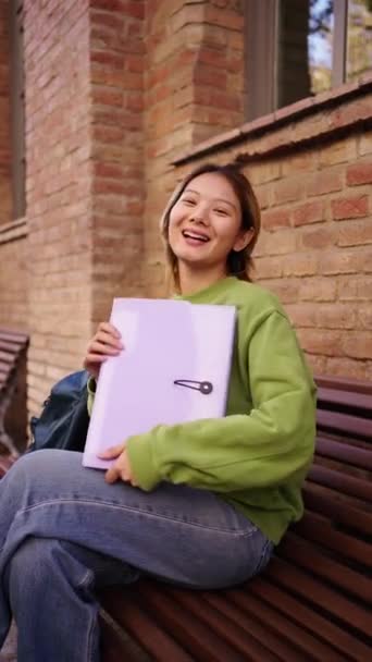 一个年轻的微笑的中国学生坐在校园的长椅上看着相机的垂直肖像 Gen Z亚洲女人手里拿着一个文件夹在大学大楼外 — 图库视频影像