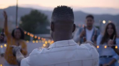 Akşamları çatıda çeşitli arkadaşlarla coşkuyla dans eden tanınmamış siyahi adamın arkasından manzara. Mutlu gençler dışarıda bira içerek bir partiyi kutluyorlar.