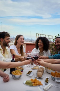 Dikey. Kahkahalar atan çeşitli genç arkadaşlar birlikte dışarıda öğle yemeği yiyorlar. Neşeli insanlar yazın kadeh kaldırarak kırmızı şarap içerek çatıdaki atıştırma partisini kutluyorlar. Mutlu yıllar.