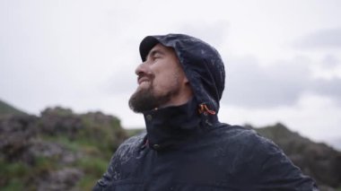 Koyu mavi Mackintosh giymiş, gülümseyen beyaz bir adam doğal ortamda tatmin olmuş görünüyor. Mutlu yakışıklı erkek doğada özgürlük ve huzur hisseder. Yağmur yağarken tatilin tadını çıkarır.
