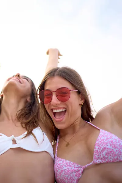 Vertical Low angle. Portrait of excited group generation z women looking camera posing on beach embracing and laughing. Cheerful female friends raising arms enjoying summer holidays in Mediterranean
