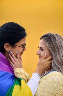 Dikey. Birbirlerine sarılıp gözlerinin içine bakan iki genç kadın. Lezbiyenler sevecen tavırlarıyla yüzlerini okşarlar. Sarı arka planda mutlu ve iyi insanlar var. Z Jenerasyonu.