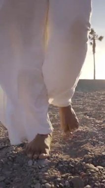 Dikey. Gün batımında beyaz pantolonlu bir kadın. Gün batımında sahilde çıplak ayakla yürürken kadın ayaklarını yavaşça kapat. Yaz tatilinde kadın turist kumsalda rahatça dolaşıyor.. 