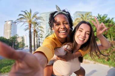 Parkta bir ağacın yanında iki mutlu kadın birlikte selfie çekerken gülümsüyor. Kolları gerilmiş, gökyüzü ve arka planda bitkiler var.