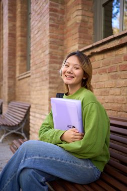Kampüste bir bankta oturmuş kameraya bakan gülümseyen genç Çinli öğrencinin dikey portresi. Asyalı bir kadın üniversite binasının dışında elinde bir dosya tutuyor.