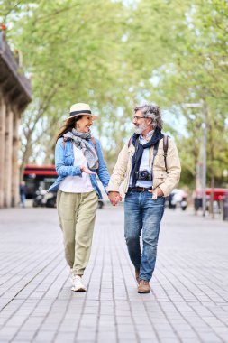 Dikey. Mutlu yetişkin Kafkas turist çifti şehir sokağı için el ele tutuşan diğer yürüyen çiftlere bakıyor. Romantik kaçamakların tadını çıkaran olgun aşk insanları. Olumlu ilişkiler ve emekli bahar tatilleri