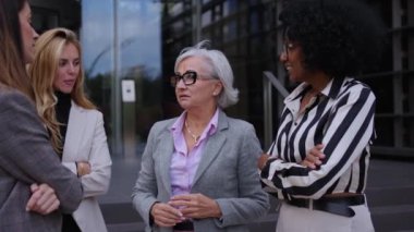 Düşük açı. İş dünyasından çeşitli yaş ve ırktan kadınlar ofis binasının dışında konuşuyorlar ve günlük toplantılar yapıyorlar. Resmi takım elbiseli profesyonel kadın girişimciler.