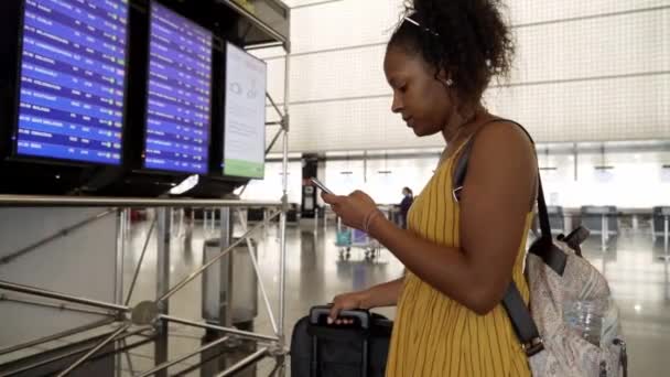 年轻的非洲裔美国女性旅行者站在机场的信息板上检查她的航班起飞时间 拉丁美洲女游客用手机到国外度假的侧视图 — 图库视频影像