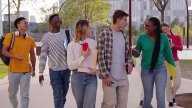 Üniversite kampüs binasının önünde birlikte yürüyen bir grup çok kültürlü öğrenci. Sırt çantaları ve çalışma kitaplarıyla gülümseyen ve birbirlerine bakan genç arkadaşlar.