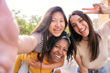 Çok ırklı bir grup genç bayan, açık havada birlikte gülümseyen selfie portresi çekiyorlar. Afrikalı bir kadın, Asyalı bir kızı sırtında taşıyor. Eğlenceli bir fotoğraf için heyecanlı güzel arkadaşlar Güneşli bir gün