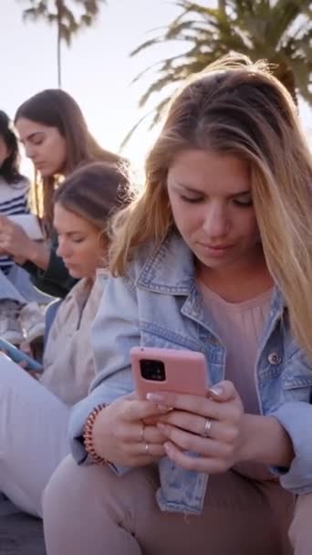 年轻的金发姑娘坐在那里 专心致志地和有背景的团体朋友一起在手机上打字 非常漂亮的女人沉迷于户外使用智能手机的技术 新世代的社会隔离 — 图库视频影像