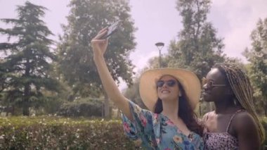 İki tane gülümseyen, etnik çeşitlilikte genç kız açık hava parkında telefonla selfie çekerken biri diğerine öpücük veriyor. Lezbiyen çift, bayram fotoğrafı için sevgiyle sarılıyor. Homoseksüel ilişkiler yaşam tarzı