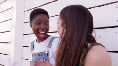 Beyaz duvara yaslanıp birlikte gülen neşeli beyaz beyaz ve Afrikalı Amerikalı kadınları yakından takip edin. İki mutlu arkadaş, güzel bayan dışarıda eğleniyor. Z kuşağındaki dostluk kavramı