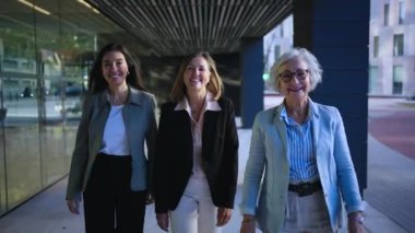 Ofis merkezinin dışındaki kamerada mutlu görünen üç iş kadını. Ön manzara sadece takım elbiseli olgun bir kadın dışarıda neşeli bir şekilde dolaşır. Pozitif güçlü profesyonel kıdemli insanlar 