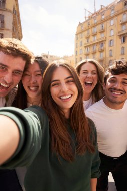 Dikey. Bir grup genç insan binanın önünde selfie çekerken gülümseyip eğleniyor. Öğrenciler turist gezileri sırasında keyifli anları paylaşıyorlar.