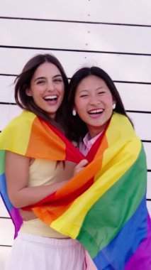 Dikey. Çok kültürlü lezbiyen çift arka tarafta sevgi dolu gökkuşağı bayrağını kucaklarken kameraya gülümsüyor. Genç Kafkas ve Asyalı neşeli kadınlar gurur günü beyaz geçmişini kutluyorlar
