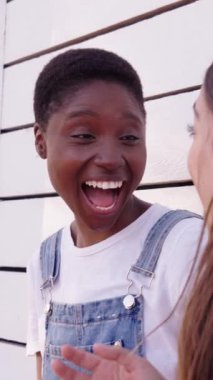 Dikey. Beyaz duvara yaslanıp birlikte gülen neşeli beyaz beyaz ve Afrikalı Amerikalı kadınları yakından takip edin. İki mutlu arkadaş, güzel bayan dışarıda eğleniyor. Z jenerasyonunda arkadaşlık