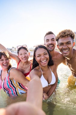 Mayo giymiş, gülümseyen ve suyun içinde kameraya bakarak selfie çeken bir grup mutlu gencin dikey hali. Denizde çok ırklı arkadaşlar plaj keyfi yapıyor ve eğleniyorlar.