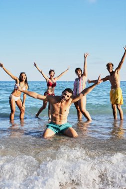 Yüzme kostümlü, sahilde fotoğraf çektirmek için poz veren mutlu gençlerin dikey portresi. Çeşitli arkadaşlar gülümsüyor ve neşeli hareketler yapıyor. Kameraya bakarak kollarını kaldırıyorlar.