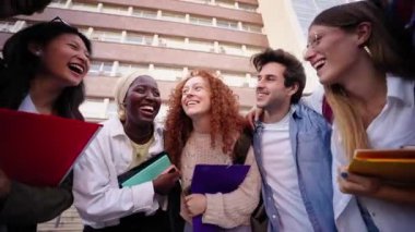 Düşük açı. Bir grup çok ırklı ve neşeli genç öğrenci dışarıda gülmekten zevk alıyor. Çeşitli arkadaşlar sırt çantaları ve çalışma kitaplarıyla kucaklaşıp ders aralarında eğleniyorlar. 