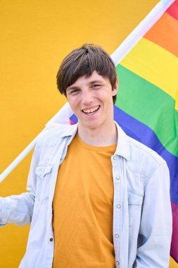 Dikey. Elinde Igbt gökkuşağı bayrağı tutan mutlu, genç bir Kafkasyalı. Eşcinsel insanlar sarı arka planda fotoğraf için poz veriyorlar. Neşeli eşcinsel insan izole edilmiş stüdyoda mutlu bir fotoğraf. Z Jenerasyonu.