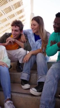 Dikey. Kalabalık mutlu, çok kültürlü gençler merdivenlerde oturmuş dışarıda birlikte komik şeylerden bahsedip gülüyorlar. Gen Z neşeli arkadaşlar iletişim ve arkadaşlık konsepti