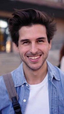 Dikey. Bulanık üniversite binasının önünde kameraya gülümseyen beyaz erkek öğrencinin portresi. Kampüsteki derslerin arasında neşeli pozlar veriyor.