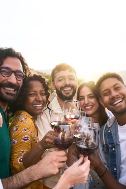 Dikey. Bir grup çok ırklı arkadaş kırmızı şarap içip kır evinde kutlama yapıyorlar. Gençler açık terasta selfie çekerken sarılıyorlar. Erkekler ve kızlar yaz tatilinin tadını çıkarıyor..