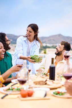 Dikey. Genç, gülümseyen, beyaz bir kadın çatıdaki mutlu insanlarla kutlamada sofraya yemek koyuyor. Bir grup neşeli çok ırklı arkadaş dışarıda barbekü partisi için toplandı.