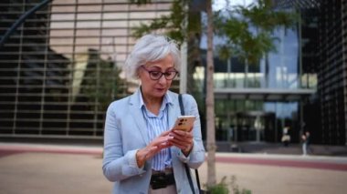 Olgun beyaz iş kadını cep telefonuyla kameraya doğru yürüyor. Kıdemli profesyonel kadın gezintiye çıkıyor. Cep telefonlarına bakıyor ve açık havada gülümsemeyi reddediyor. Ofise giden insanlar.