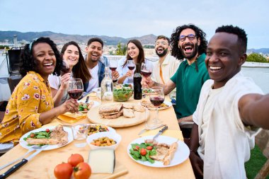 Genç Afrikalı gülümseyen adam barbekü masasında oturan neşeli milenyum insanlarına selfie çekiyor. Yazın çatıdaki toplantının portresi. Dışarıdaki kameraya bakarak mutlu poz veren çok ırklı arkadaşlar. 