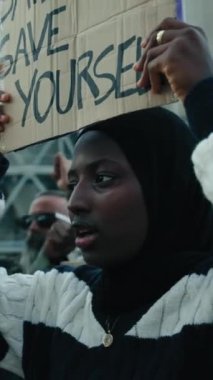 Dikey video. Ciddi Müslüman bir kadın, iklim değişikliği ve küresel ısınma, kirlilik ve fabrikalara karşı düzenlenen bir gösteride bir grup insanın protesto pankartı tutuyor.