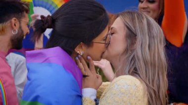 İki çok ırklı genç romantik kadın dışarıda gay onur festivalinde öpüşüyor. Arka planda LGBT partisini gökkuşağı bayrağı ve hayranlarıyla kutlayan bir grup arkadaş. Z jenerasyonu ve cinsel özgürlük. 