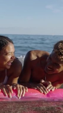 Mayo giymiş, gülümseyen ve şişme bir plaj minderinde eğlenen bir grup mutlu gencin dikey görüntüsü. Denizde çok ırklı arkadaşlar plaj keyfi yapıyor ve eğleniyorlar.