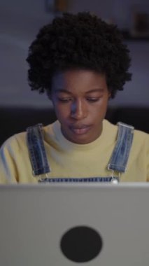 Dikey. Gece odasında dizüstü bilgisayar kullanan serbest çalışan genç bir afro kadın. Afrika kökenli Amerikalı kadın bilgisayar ekranına bakıyor. Milenyum Siyahi Kişi Evden İnternet 'te Çalışıyor