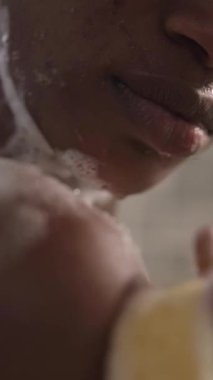 Dikey. Afro-Amerikalı bir kadın, duşta omuzlarından yüzüne kadar rahatlayarak derisini temizliyor. Siyah yetişkin bir kadının günlük temizlik için sabun kullanması. Evdeki tuvalette kişisel vücut bakımı