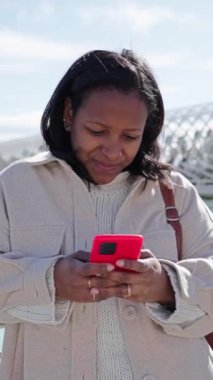 Dikey. Gülümseyen Latin kadın Valencia 'daki bilim parkında cep telefonu kullanıyor ve bakıyor. Güneşli kış gününde hücrede yazı yazan zenci bir turist. Seyahatte iletişim kurmak