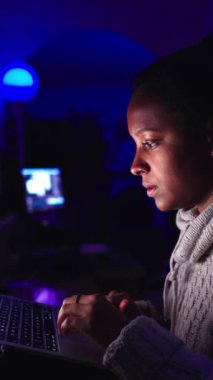 Dikey. Genç siyah kadın karanlıkta dizüstü bilgisayarda klavye kullanıyor. Merkez ofiste neon ışıklarıyla çalışan konsantre Afro-Amerikan kadın. İş, telekomünikasyon, programlama ve serbest.