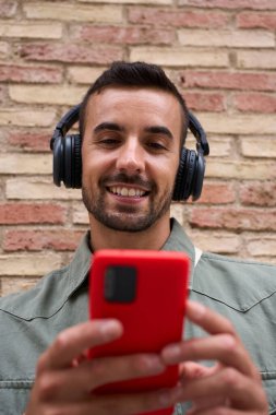 Duvara yaslanmış kırmızı cep telefonu kullanan çekici bir delikanlının dikey yakın çekimi. Milenyum Avrupalı erkek açık havada durup gülümseyerek gezmekten zevk alıyor. Teknolojiye bağımlı insanlar 