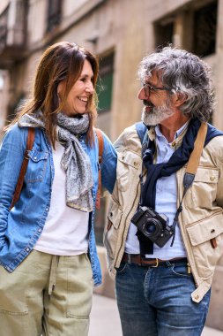 Vertical. Cheerful senior attractive couple look at each other smiling on tourist street. Retirees enjoying leisure free time together on vacation outdoors. Mature marital relationship and pensioners
