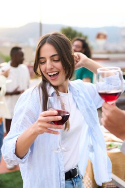 Dikey. Çatıdaki partide arka planda arkadaşlarıyla dans eden mutlu, çok kültürlü bir çift. Beyaz kadın gün batımında barbeküde kırmızı şarap içerek eğleniyor. İnsanlar yaz tatilini kutluyor.