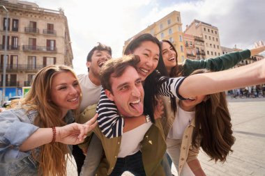 Bir grup mutlu genç bir binanın önünde poz veriyor, gülümsüyor ve jestler yapıyor. Boş zamanlarının tadını çıkarıyorlar, bulutlu bir gökyüzü altında seyahat deneyimlerini paylaşıyorlar.