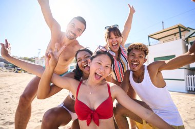 Sahilde eğlenen, gülümseyen ve kollarını kaldıran neşeli, neşeli, çeşitli genç arkadaşların komik bir portresi. Yaz tatilinde mayo giyen Z jenerasyonu insanları