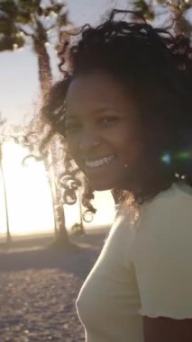 Dikey. Afro-Amerikalı kadın kumsalda rahatça yürüyüp gülümseyen kameraya bakıyor. Yaz tatilinde günbatımının tadını çıkaran genç afro bir kadın. Tatil yeri ve mutlu insanlar..