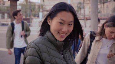 Üniversite arkadaşlarıyla gülümseyen kameraya bakan genç Asyalı kadın şehir meydanında tatilin tadını çıkarıyor. Mutlu Çinli gen z kadın arka planda çeşitli insanlarla poz veriyor.