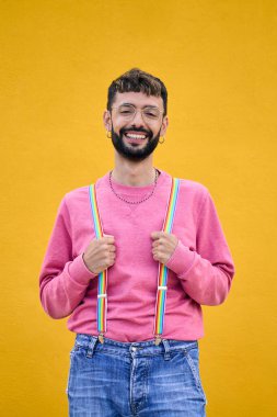 Dikey. Portre gülümseyen genç beyaz adam gökkuşağı lgbt pantolon askısını tutuyor. Sarı arka planda fotoğraf için poz veren eşcinseller. Neşeli erkek sakallı insan izole edilmiş stüdyoda mutlu..